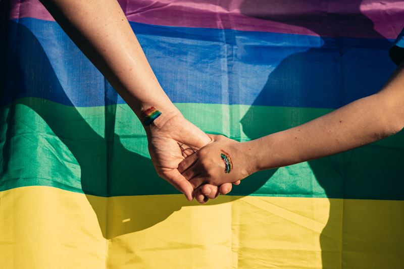 A close up of two hands holding in front of a pride flag. There are rainbows painted on their hands